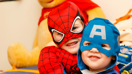 A boy wearing a spiderman mask and a boy wearing a captain america mask hug while smiling at the camera.